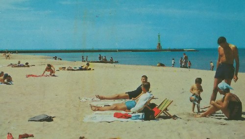 A sunny beach scene with people sunbathing, playing, and a lighthouse in the distance.