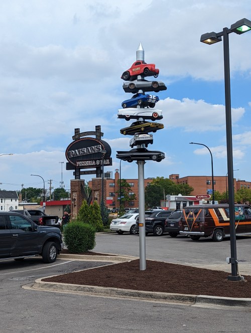 A tall pole displays stacked toy cars outside a pizzeria, with a sign for the restaurant in the background.