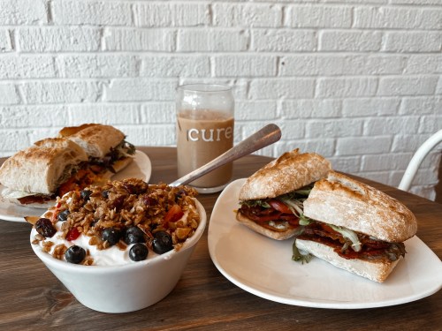 A breakfast spread featuring a yogurt bowl with granola and berries, a sandwich, and a drink on a wooden table.