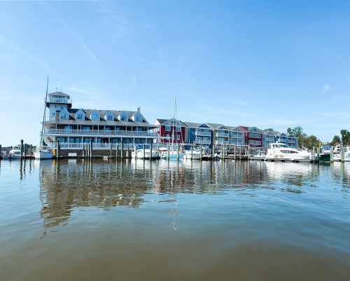 A marina with colorful buildings and boats reflected in calm water under a clear blue sky.