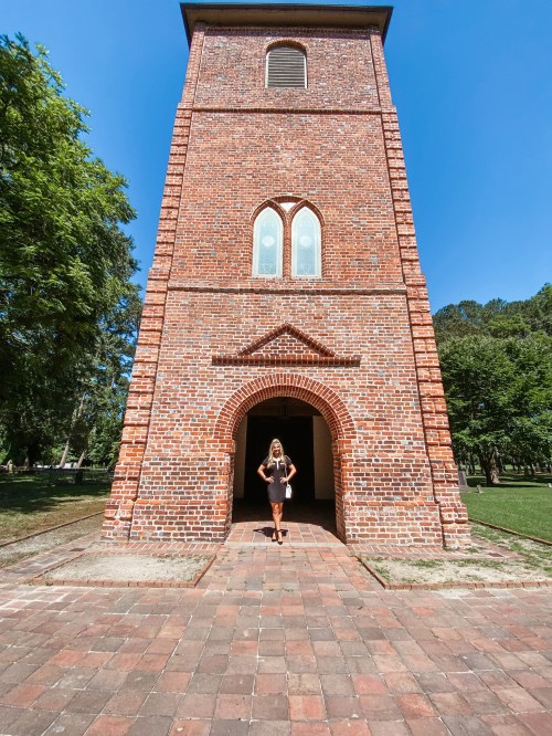 A woman stands in front of a tall brick tower with arched windows, surrounded by greenery and a clear blue sky.