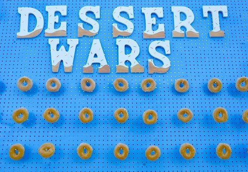 Colorful letters spelling "DESSERT WARS" above a display of donuts on a blue pegboard background.
