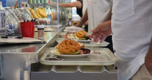A person holds a tray with food, including a burger and onion rings, in a cafeteria setting.