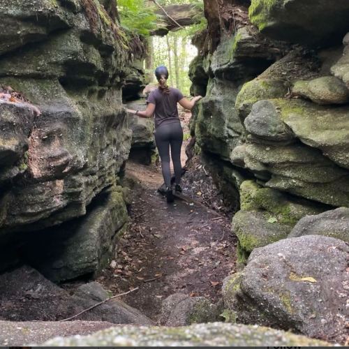 A person walking through a narrow rock formation in a forested area, surrounded by lush greenery.