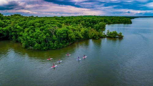 Aerial view of kayakers paddling on a calm river surrounded by lush green trees and a cloudy sky.