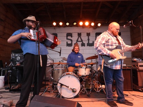 A band performs on stage with an accordionist, guitarist, and drummer, under warm stage lights.