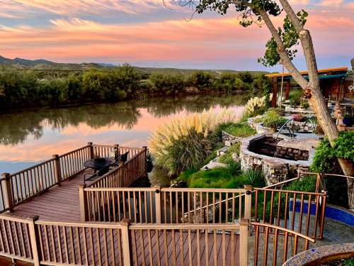 Scenic view of a river at sunset, with a wooden deck, lush greenery, and a cozy outdoor seating area.