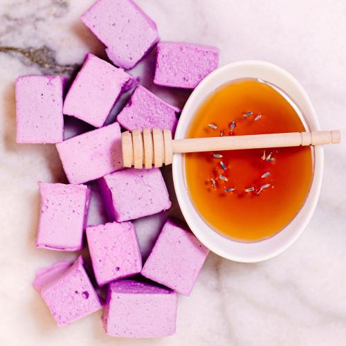 A bowl of honey with lavender sits beside purple marshmallows on a marble surface.