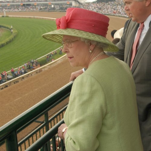 A woman in a pink hat and glasses watches a horse race from a balcony, with a crowd and racetrack in the background.