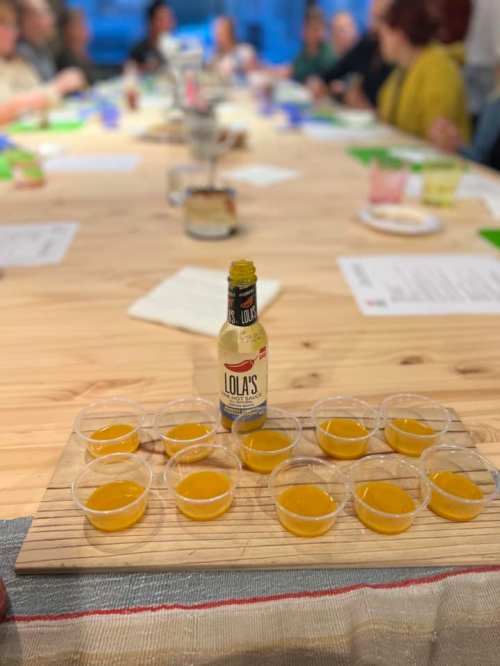 A wooden table with small cups of yellow liquid arranged in a grid, featuring a bottle of Lola's in the center.