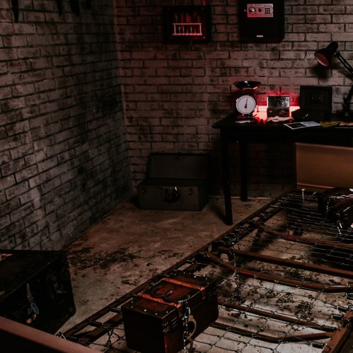 A dimly lit room with a bed frame, scattered tools, a desk with a clock, and various storage boxes against brick walls.