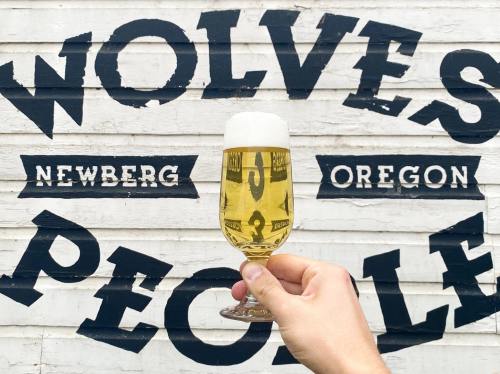 A hand holds a glass of beer in front of a mural that reads "Wolves People Newberg Oregon."