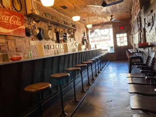 A rustic bar interior with wooden stools, vintage decor, and a door leading outside. Warm lighting enhances the atmosphere.
