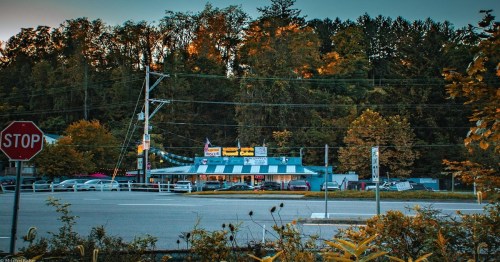 A roadside view of a diner with a striped awning, surrounded by autumn trees and a stop sign in the foreground.