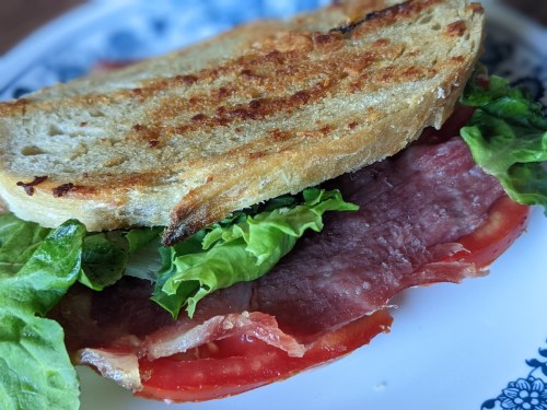A close-up of a sandwich with toasted bread, layers of lettuce, tomato, and sliced meat on a decorative plate.