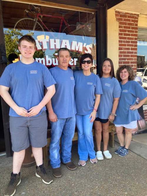 A group of five people in matching blue shirts stand outside a burger restaurant, smiling at the camera.