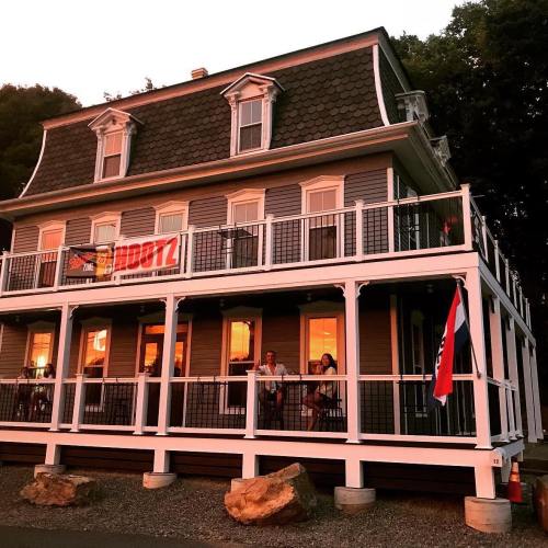 A large, two-story house with a wraparound porch, featuring a banner and people sitting outside at sunset.