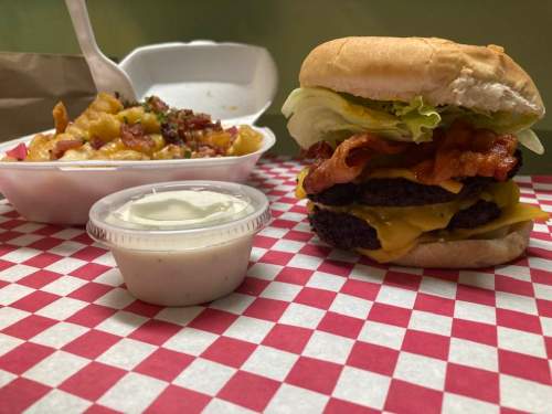 A cheeseburger with bacon and lettuce next to a container of loaded mac and cheese on a checkered tablecloth.