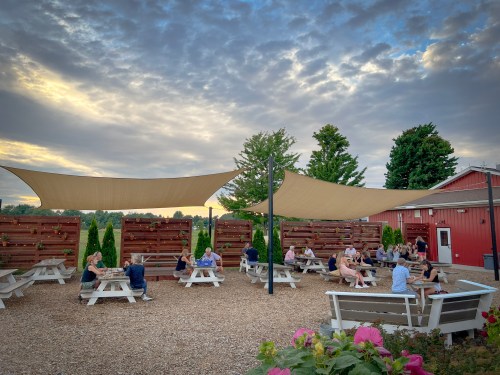 Outdoor seating area with shaded tables, people enjoying drinks, and a red barn in the background under a cloudy sky.