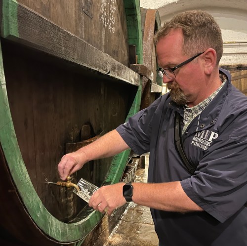 A man fills a glass from a large wooden barrel in a historic setting.