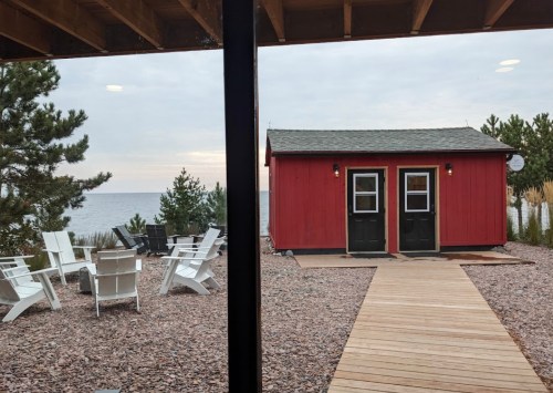 View of a red cabin by the water, with a wooden path and outdoor seating area surrounded by trees.