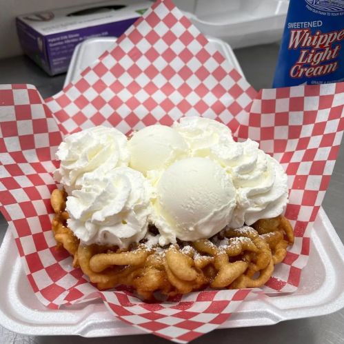 A funnel cake topped with three scoops of ice cream and whipped cream, served in a checkered paper basket.