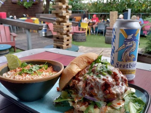 A burger with toppings, a bowl of rice, and a can of beer on a table, with a vibrant outdoor setting in the background.