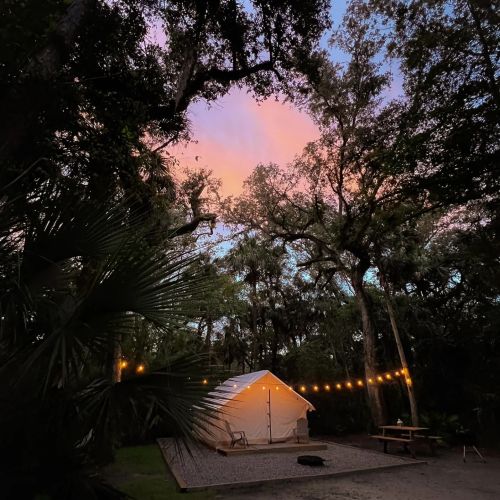 A cozy tent surrounded by trees, illuminated by string lights, with a colorful sunset sky in the background.