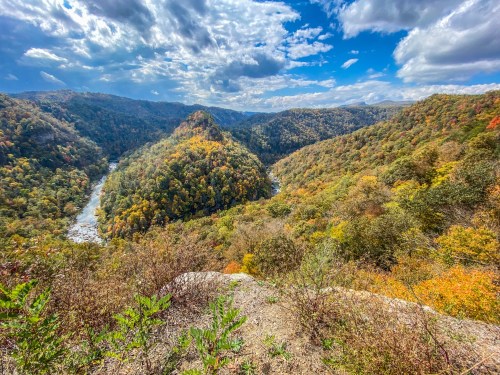 A scenic view of a river winding through colorful autumn foliage in a mountainous landscape under a partly cloudy sky.