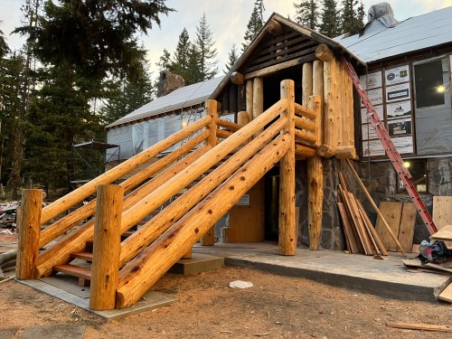 A wooden structure with log pillars and a ramp, under construction near a building surrounded by trees.