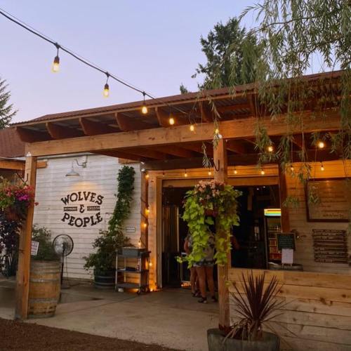 A cozy outdoor venue with string lights, a sign reading "Wolves & People," and lush greenery at dusk.