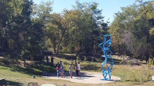 A group of people gathers around a blue abstract sculpture in a park with trees and greenery in the background.