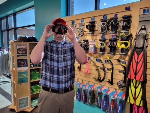 A person in a plaid shirt and red cap holds snorkeling masks in a store with various water sports gear displayed.