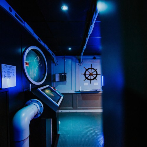 A dimly lit room with a ship's wheel, control panel, and a large circular gauge on the wall, all in blue tones.