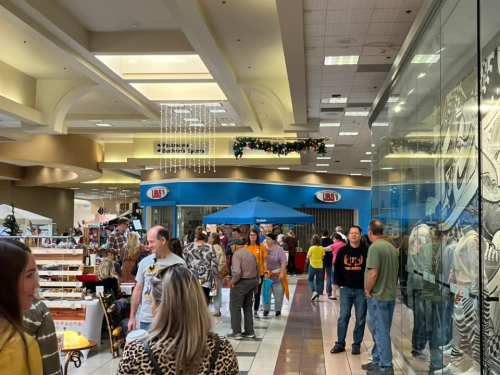 A busy mall corridor with shoppers, booths, and a blue tent, decorated for the holidays.