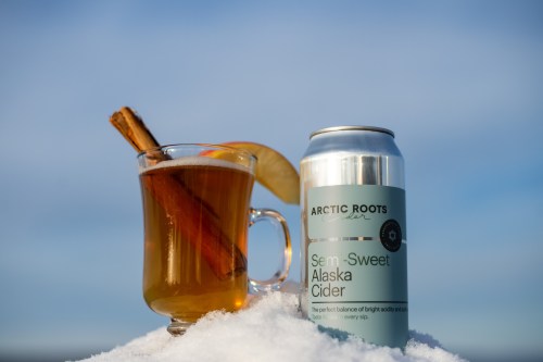 A glass of warm cider with a cinnamon stick and apple slice beside a can of Arctic Roots Semi-Sweet Alaska Cider on snow.