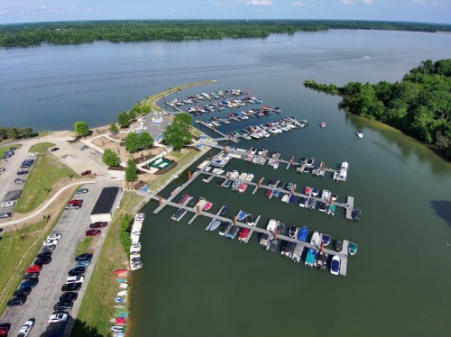 Aerial view of a marina with numerous boats docked, surrounded by green trees and a calm lake.
