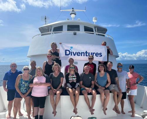 Group photo of people on a boat holding a "Diventures" banner, with blue skies and ocean in the background.