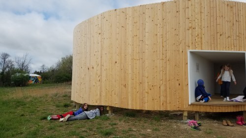 Two children sit on the ground near a circular wooden structure, while another child plays inside. Green grass surrounds them.