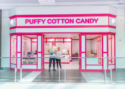 A man and a woman stand in front of a bright pink cotton candy shop with the sign "Puffy Cotton Candy."