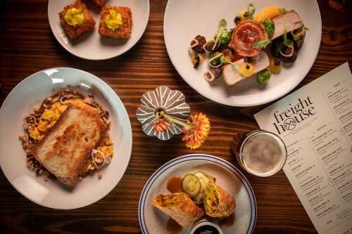 A top-down view of a table with various dishes, including a sandwich, salad, and appetizers, alongside a menu and flower.