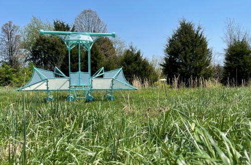 A teal playground structure sits in a grassy field under a clear blue sky, surrounded by trees.