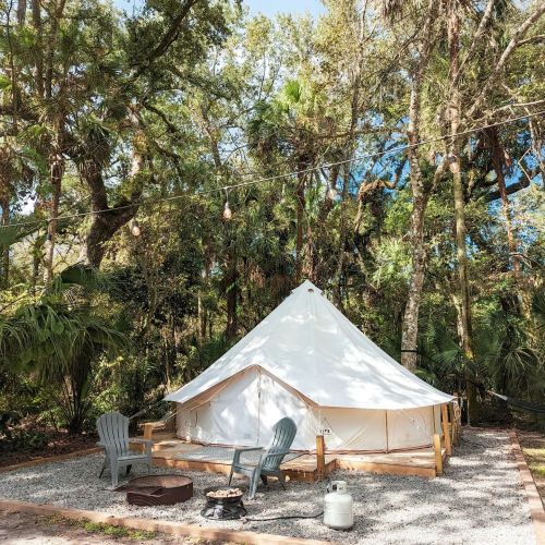 A white glamping tent surrounded by trees, with two chairs and a fire pit in front on a gravel area.