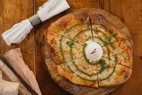 A wooden platter holds a sliced pizza topped with cheese, green herbs, and a dollop of cream, on a rustic table.