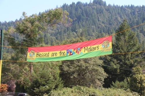 A colorful banner reads "Blessed are the Peace Makers" against a backdrop of green trees and mountains.
