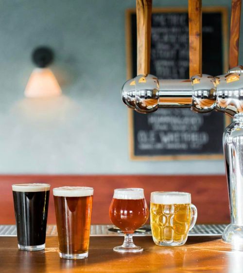 A bar counter with four glasses of different beers in front of a shiny tap and a chalkboard in the background.