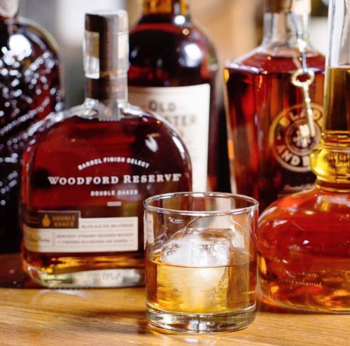 A selection of whiskey bottles with a glass of whiskey on the rocks in the foreground.
