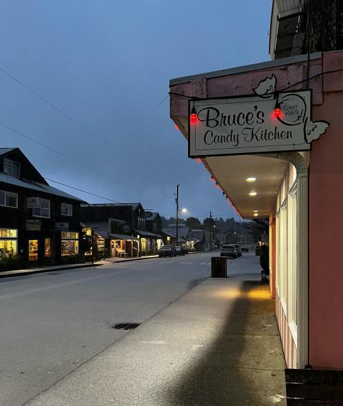 A quiet street at dusk featuring Bruce's Candy Kitchen with festive lights and nearby shops.