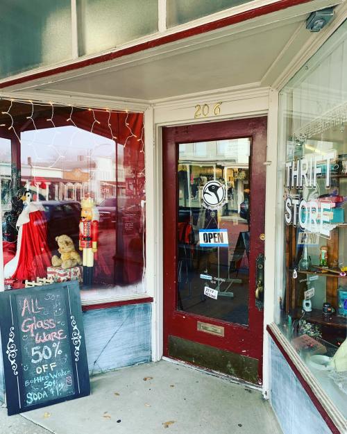 Entrance of a thrift store with a red door, holiday decorations, and a sign advertising a glassware sale.
