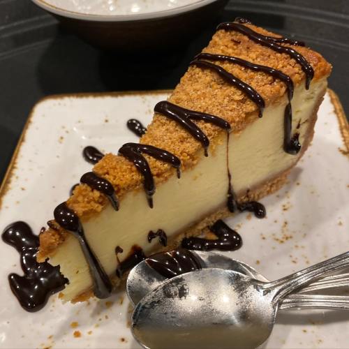 A slice of cheesecake with a graham cracker crust, drizzled with chocolate sauce, on a white plate with two spoons.